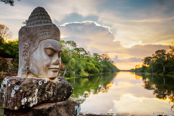 Stone face Asura on causeway near South Gate of Angkor Thom in Siem Reap, Cambodia. Beautiful sunset over ancient moat in background. Mysterious Angkor Thom is a popular tourist attraction.