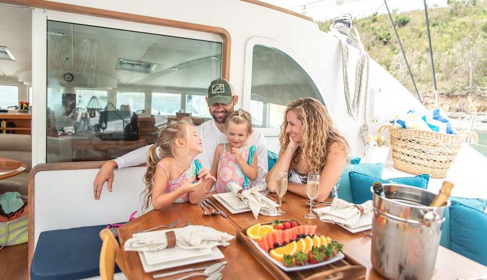 Family w 2 girls eating fresh fruit and drinking champagne aboard 57ft Lagoon catamaran TABULA RASA. Yachting with youngsters and teens.