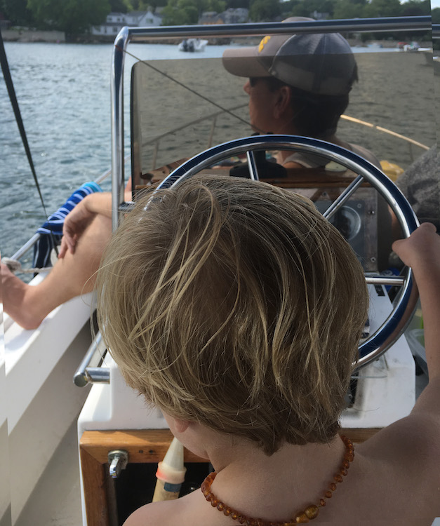 Boy at the helm of boat with his Dad. Photo by Carol Kent. Yachting with youngsters and teens.