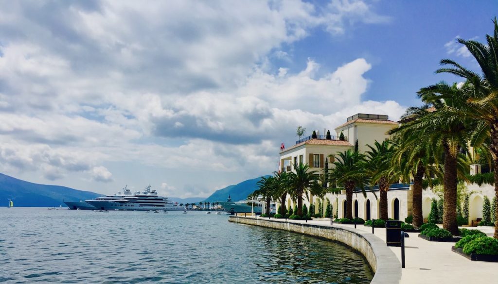 Palm tree-lined promenade with arched building and two super yachts at the marina near mountains in Kotor, Montenegro. Photo©carolkent.com